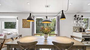 Uma área de refeições com uma mesa de madeira, quatro cadeiras e um vaso de flores. Há luminárias pendentes acima da mesa, e uma cozinha com eletrodomésticos é visível ao fundo.