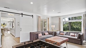 Uma sala de estar moderna com um sofá, um banquinho e uma mesa de pebolim. Ao fundo, há uma área de jantar com mesa e cadeiras.