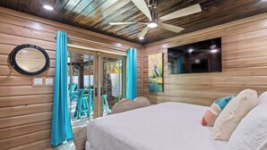 Un dormitorio con cama, ventilador de techo, televisor y puerta corrediza de vidrio que da a un área exterior con sillas y una mesa.