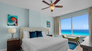 A hotel room with a large bed, a ceiling fan, artwork on the walls, a view of the ocean, and a desk with a chair.