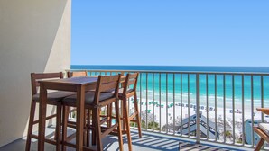 A balcony with a wooden table and chairs, overlooking a beach with umbrellas and a staircase leading down to the sand.