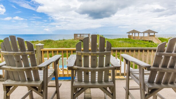 Drie Adirondack-stoelen op een houten terras met uitzicht op een strand, met een houten constructie op de achtergrond.