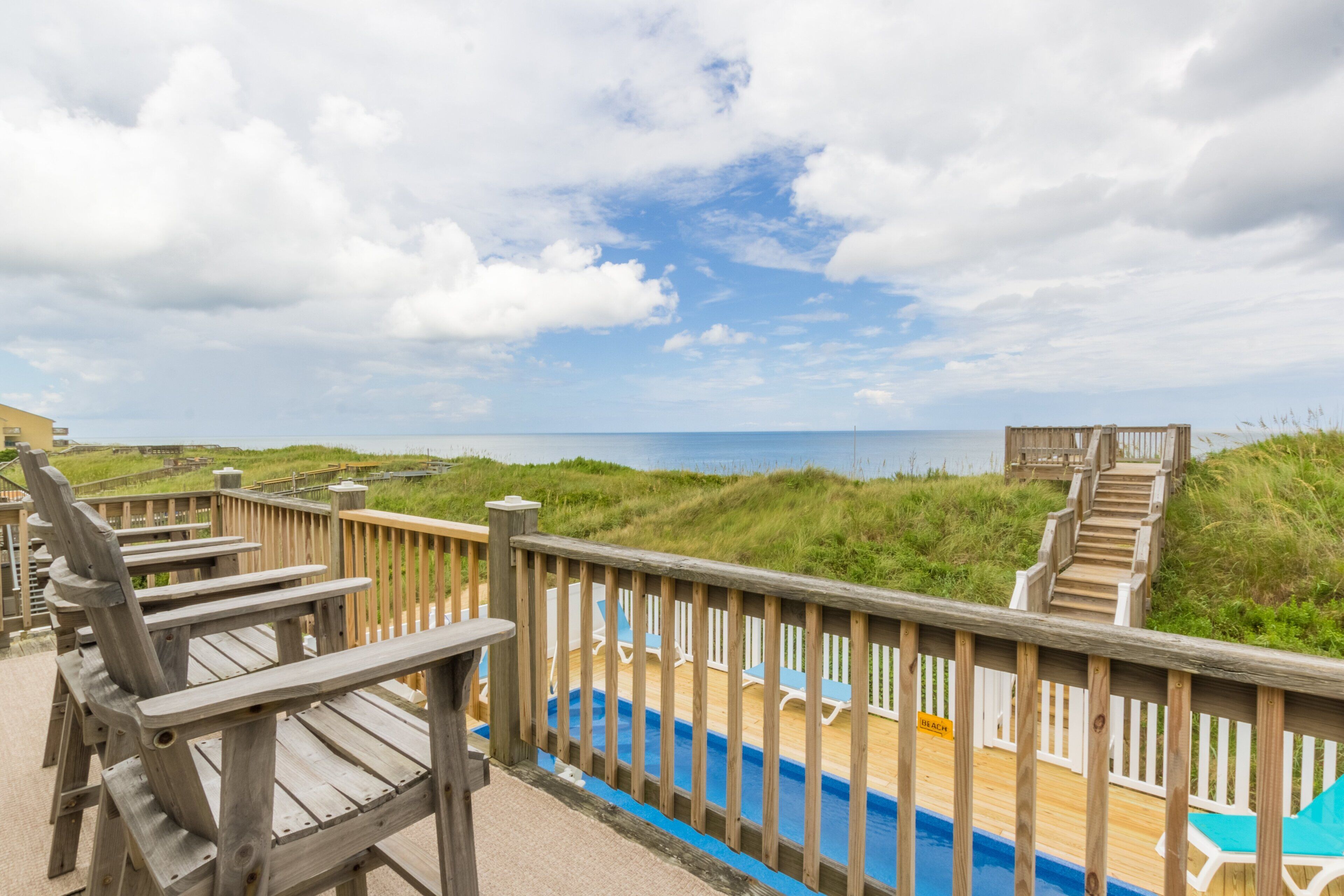 Une terrasse en bois avec une piscine, des chaises longues et une vue sur la plage et l’océan.