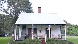 A white building with a red roof, a brick chimney, and a porch with wooden chairs.