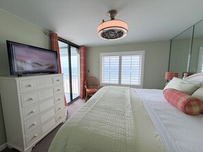 Une chambre à coucher comprenant un lit, une commode, un téléviseur, une chaise, une fenêtre avec des stores et un ventilateur de plafond.