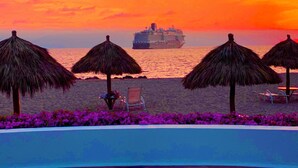 Escena de playa con un crucero a lo lejos, sombrillas de paja y una zona de piscina.