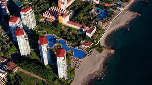 Vista aérea de un complejo turístico costero con varios edificios altos, una playa y una piscina grande.
