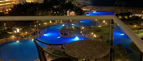 A nighttime view of a resort with a pool area, including a lounge chair and table, and a view of the ocean and a distant illuminated building.