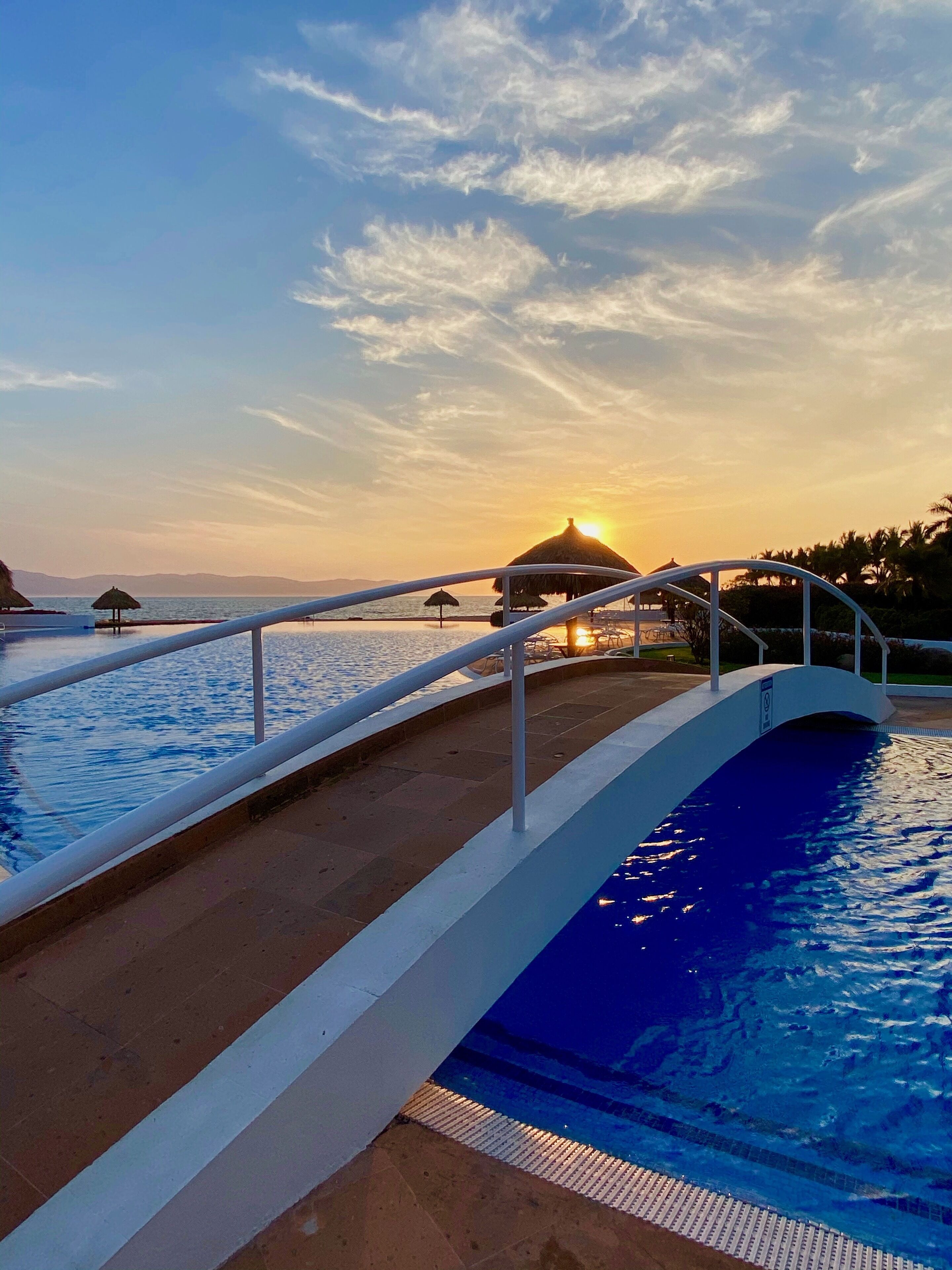A swimming pool with a white deck and a railing, a sunset in the background, and a thatched-roof structure.