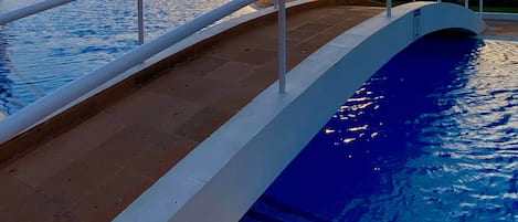 A swimming pool with a white deck and a railing, a sunset in the background, and a thatched-roof structure.