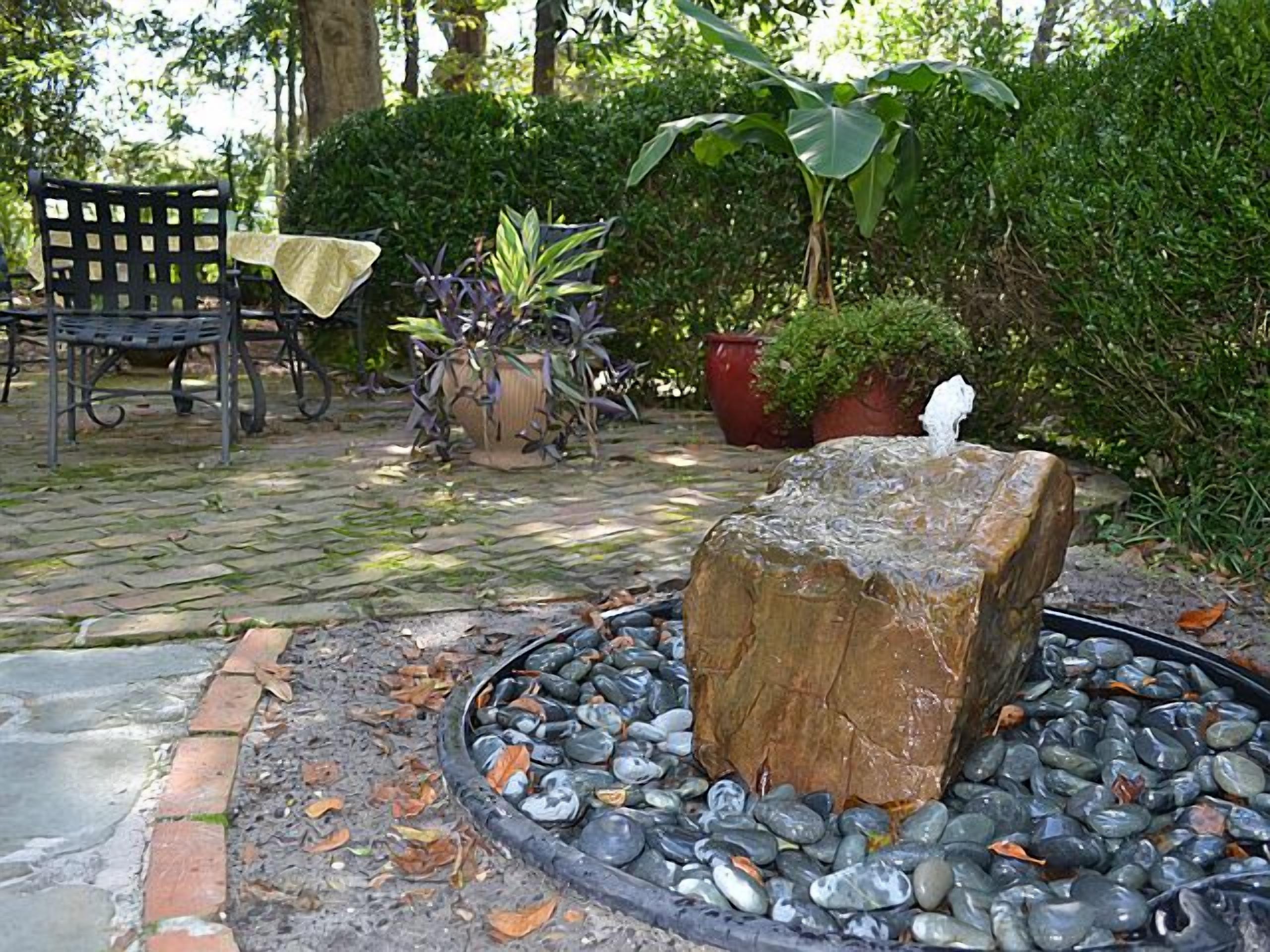 A garden area with a stone fountain surrounded by pebbles, a wooden bench, a table with a tablecloth, and various potted plants.
