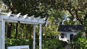 A white pergola with a bench, surrounded by lush greenery and a stone pathway leading to a house.