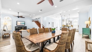 A spacious dining area with a wooden table and wicker chairs, a staircase, and a kitchen in the background.
