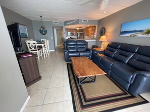 A living room with a sofa, a coffee table, and a TV. There is a kitchen in the background.