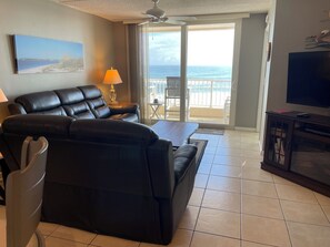 A living room with a black leather sofa, a coffee table, a TV stand, and a balcony with a view of the ocean.