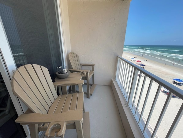 A balcony with two Adirondack chairs, a small table, and a bowl. The balcony overlooks a beach with umbrellas and people.