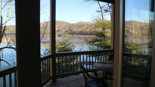 A wooden deck with a table and chairs, overlooking a lake surrounded by trees and hills.