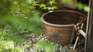 A rustic outdoor setting with a large metal container, surrounded by rocks and greenery. - Sadasuke (Kami)