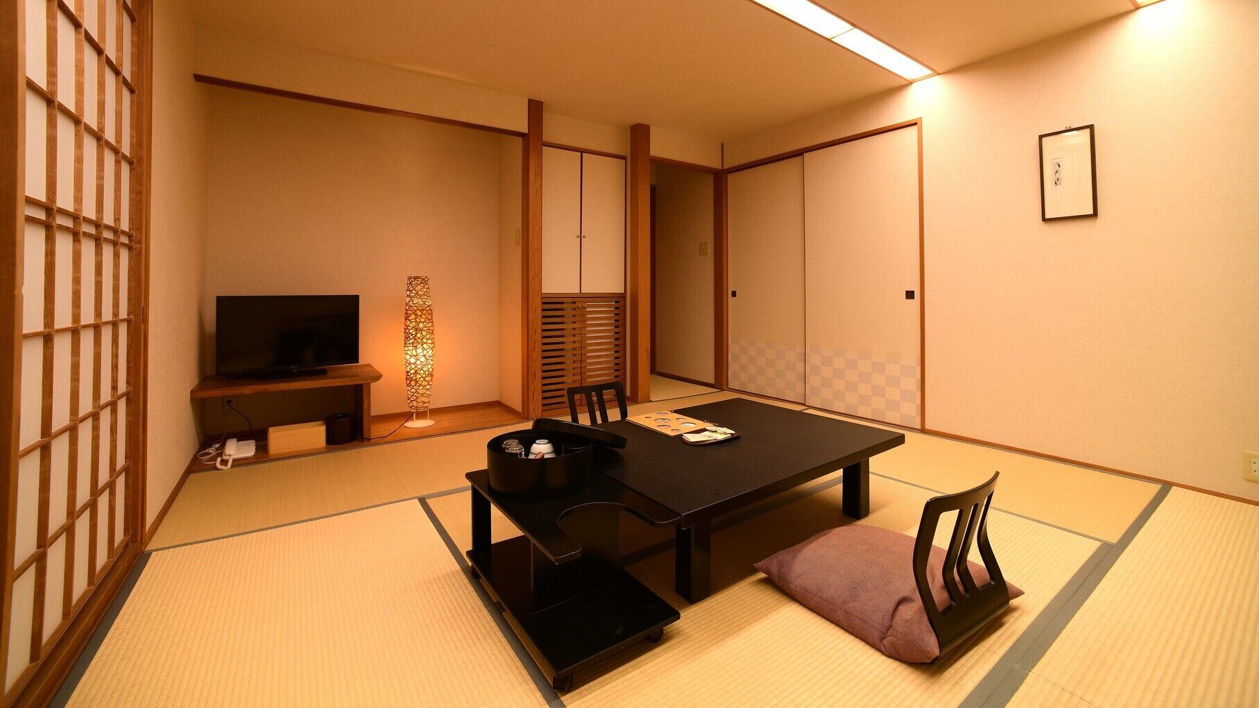 Ein traditioneller japanischer Raum mit Tatami-Böden, einem niedrigen Tisch, einem Fernseher und einer dekorativen Lampe.