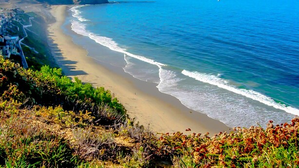 A coastal landscape with a sandy beach, a cliff, and a clear blue sky.