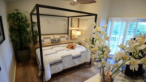A four-poster bed with a wooden frame, a bedside table with a lamp, a potted plant, and a window with a view of greenery.