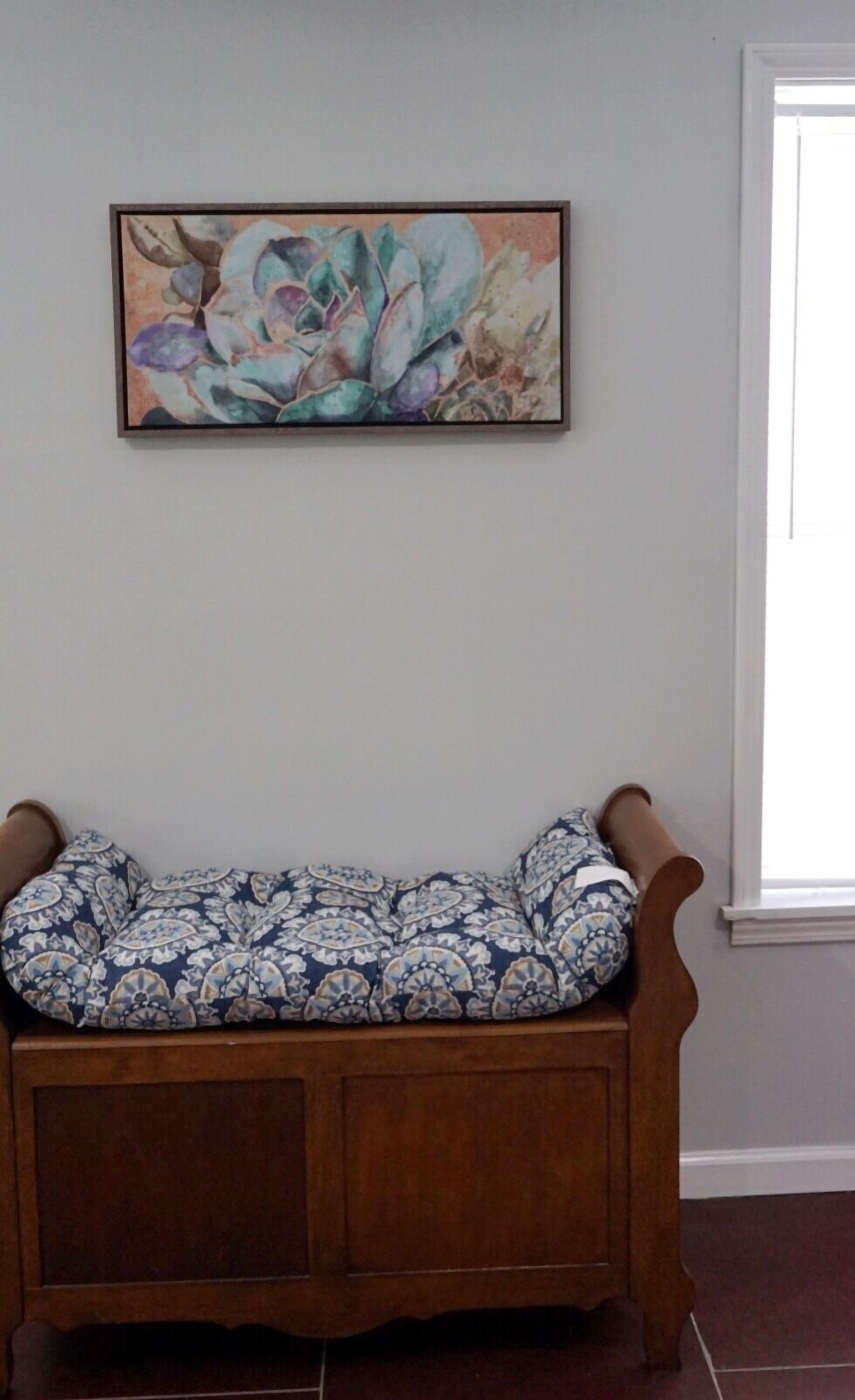 A wooden bench with a patterned cushion in a room with a plain wall and a window.