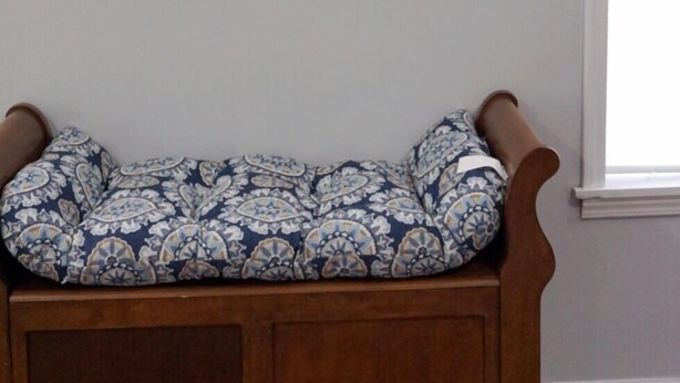Un banco de madera con un cojín estampado en una habitación con una pared lisa y una ventana.