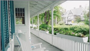 A covered porch with white railings and benches, overlooking a garden and houses. - Historic Petunia Cottage: Large Renovated Victorian Excellent In-town Location (Bar Harbor)