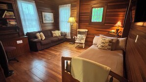 A cozy log cabin room with a wooden floor, a bed, a sofa, a rocking chair, and a desk with a lamp. - Historic Petunia Cottage: Large Renovated Victorian Excellent In-town Location (Bar Harbor)