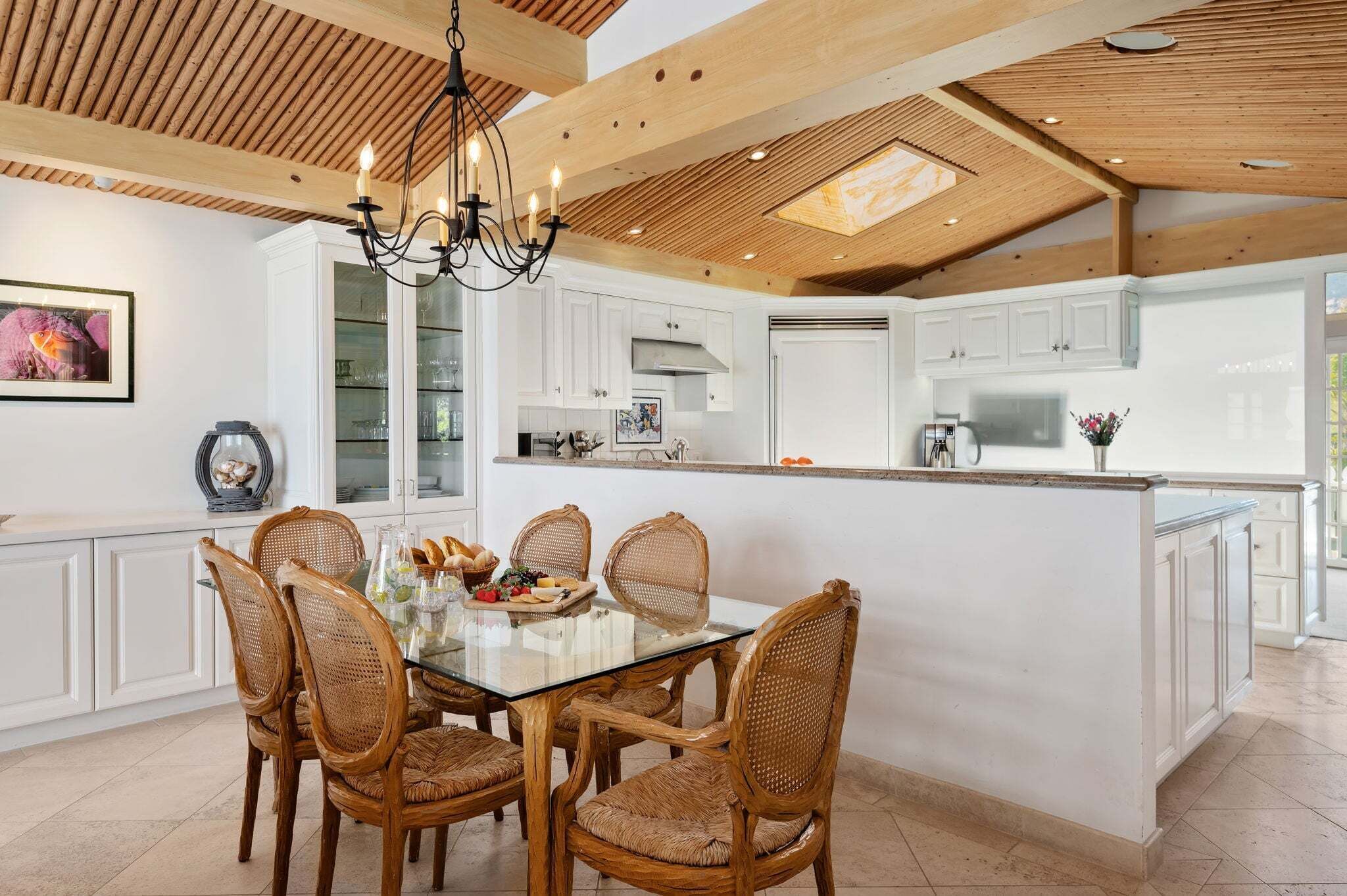 Una cocina moderna con armarios blancos, una mesa de comedor de vidrio, sillas de mimbre y una araña.