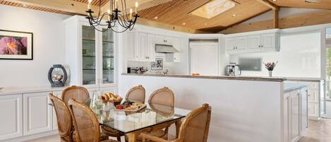 Una cocina moderna con armarios blancos, una mesa de comedor de vidrio, sillas de mimbre y una araña.