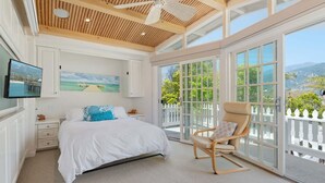 A bedroom with a bed, a TV, a painting, a chair, and a balcony with a view. - Beautiful Oceanfront Home with Jacuzzi (Carpinteria)