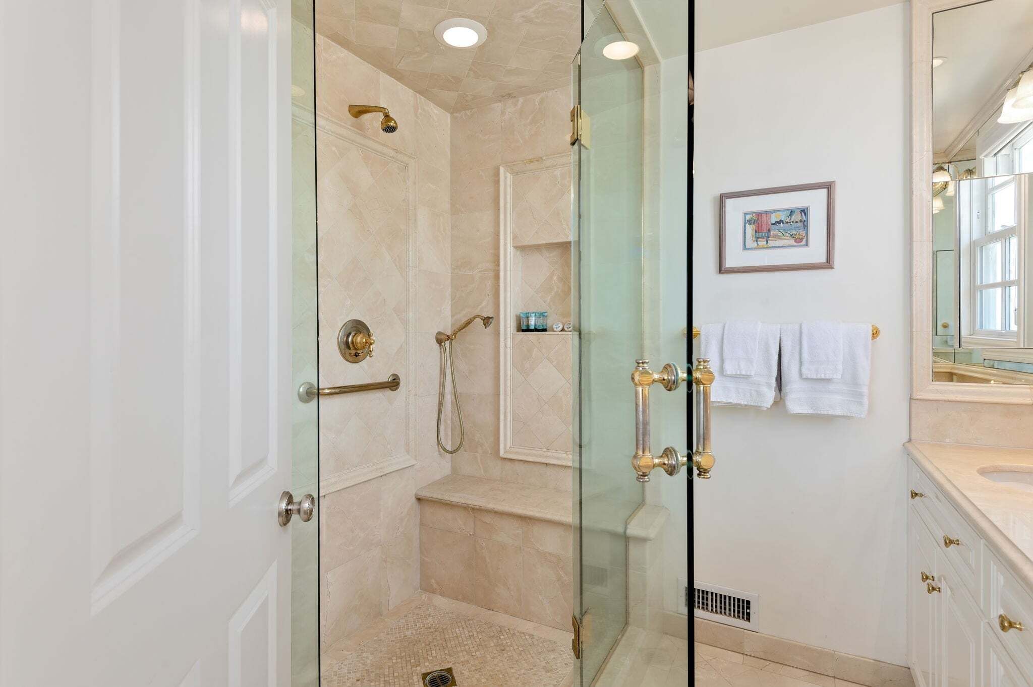 Une salle de bain dotée d’une douche à parois de verre, d’un meuble-lavabo blanc avec des accessoires dorés et d’un tableau encadré au mur.
