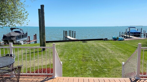 Eine Holzterrasse mit Tisch und Stühlen, eine Rasenfläche und Blick auf das Wasser und den Steg.