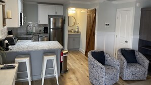 Eine moderne Küche mit weißen Schränken, einem Edelstahlkühlschrank und einer Arbeitsplatte aus Granit. Zwei weiße Barhocker und ein gemustertes Sessel sind ebenfalls vorhanden.