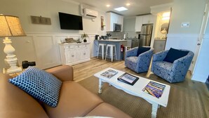Ein Wohnzimmer mit einer Couch, einem Couchtisch und gemusterten Sesseln. Im Hintergrund ist eine Küche mit weißen Schränken, einer Mikrowelle und einem Kühlschrank zu sehen.