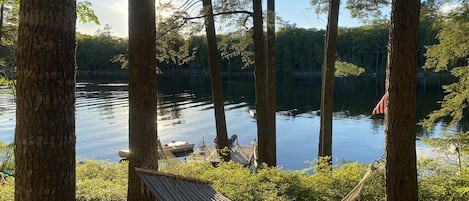A hammock between trees by a calm lake with a dock and a flag.