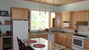 A kitchen with wooden cabinets, a refrigerator, a microwave, an oven, and a dining area with a table and chairs. - PRIVATE LUXURY, Beachfront Home, Hot Tub, Kayaks, Paddle Board, Cabana (Casuarina Point)