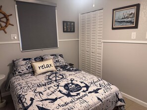 Een slaapkamer met een sprei met een nautisch thema, een 'relax' kussen, een decoratie van een scheepsschroef en een ingelijste foto van een haven.