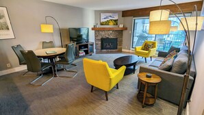 A living room with a stone fireplace, a television, a dining table with chairs, a yellow armchair, a sofa, and a floor lamp. - Luxury Condo w/ FREE WiFi, Parking, Heated Pool, Hot Tubs, Skier Shuttles (Avon)