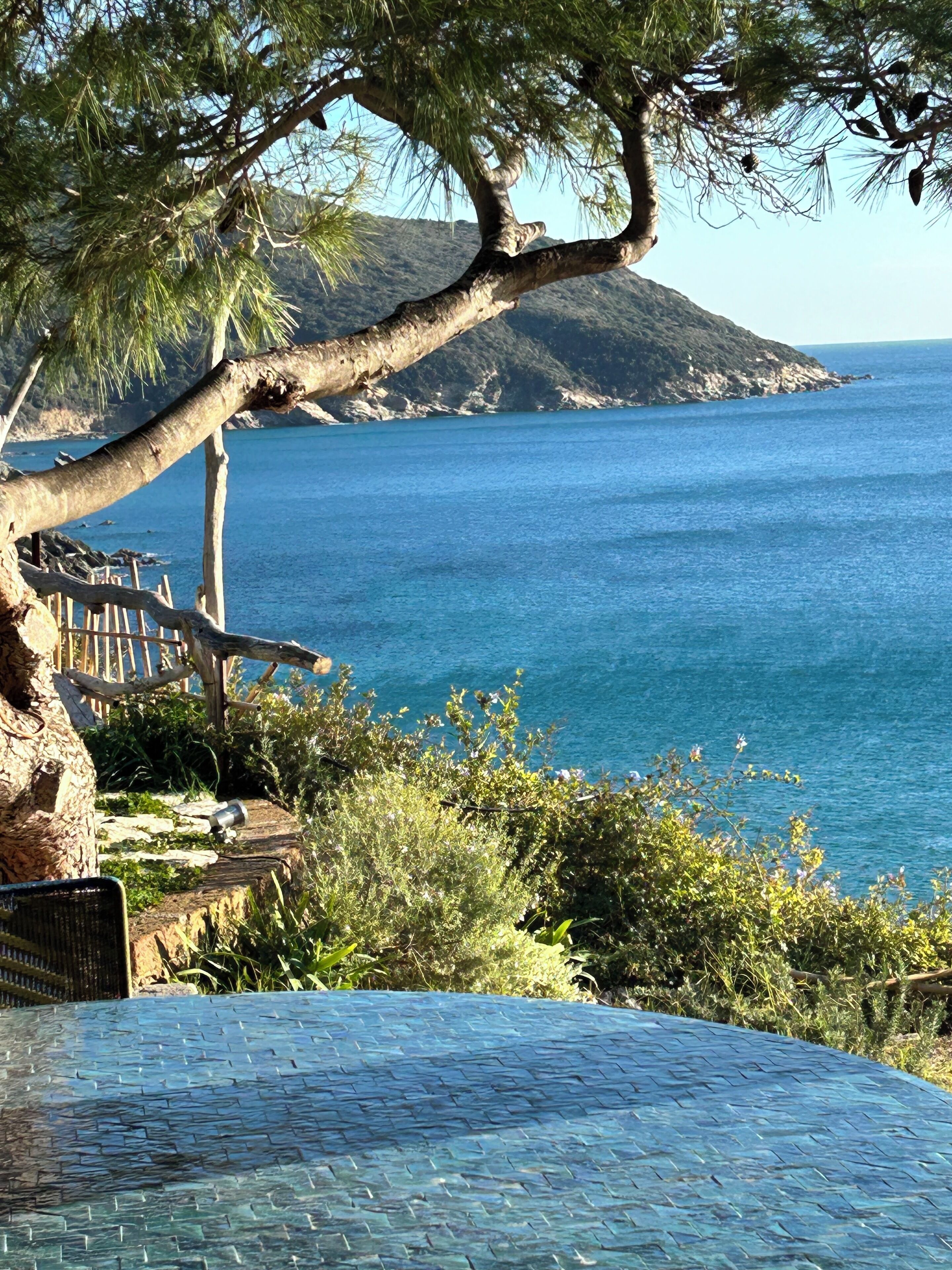 A coastal view with a clear blue sea, a mountainous backdrop, and a wooden bench overlooking the water.
