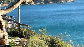 A coastal view with a clear blue sea, a mountainous backdrop, and a wooden bench overlooking the water.