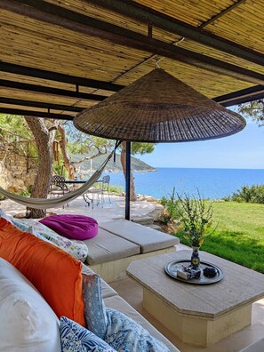 A covered outdoor seating area with a view of the sea, a hammock, and a table with a vase of flowers.