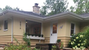 A two-story house with a stone chimney, a front porch with a bench, and a well-maintained garden with various shrubs and flowers. - Peaceful, Cozy, Clean, Fun Mid-Century Stay! Backyard Oasis Waterfall, Fireplace (Fish Creek)