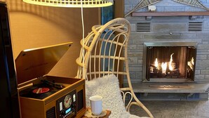 A cozy living room with a fireplace, a wicker chair, a table with a vintage radio, and a decorative hanging light. - Peaceful, Cozy, Clean, Fun Mid-Century Stay! Backyard Oasis Waterfall, Fireplace (Fish Creek)