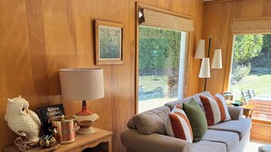 A cozy living room with a wooden wall, a grey sofa with colorful cushions, a wooden side table with a lamp, and a window with a view of greenery. - Peaceful, Cozy, Clean, Fun Mid-Century Stay! Backyard Oasis Waterfall, Fireplace (Fish Creek)