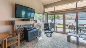 A living room with a flat-screen TV, a fireplace, and a view of a lake through large sliding glass doors.