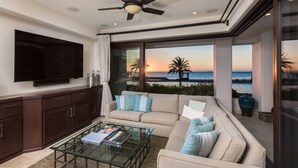 Ein modernes Wohnzimmer mit einem großen Flachbildfernseher, einem Glastisch und einer Couch mit vielen Kissen. Das Zimmer verfügt über einen Deckenventilator, große Fenster mit Blick auf den Ozean und Palmen sowie eine dunkle Holzkommode.