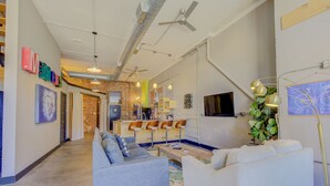 Una sala moderna con un sofá gris, un sillón blanco y una mesa de centro. Hay dos ventiladores de techo, una zona de bar con taburetes y una planta grande.