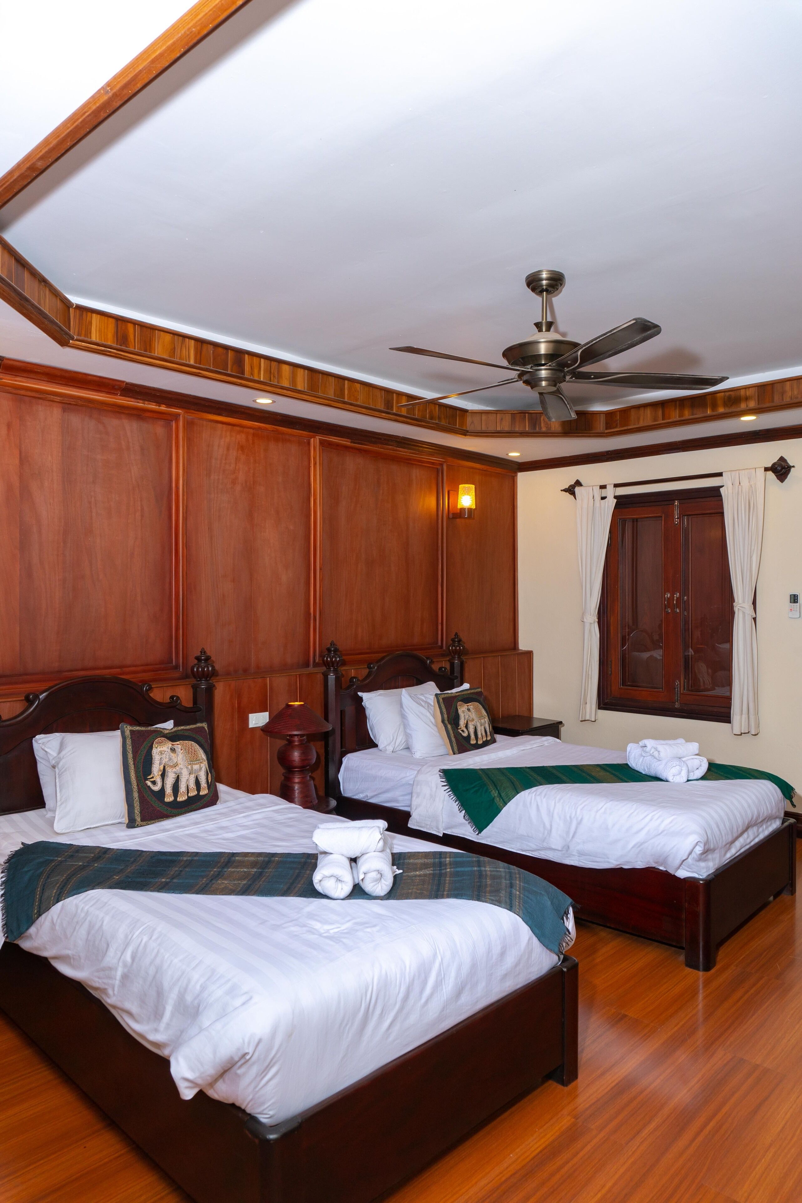 A hotel room with two beds, wooden paneling, a ceiling fan, and a window with curtains.